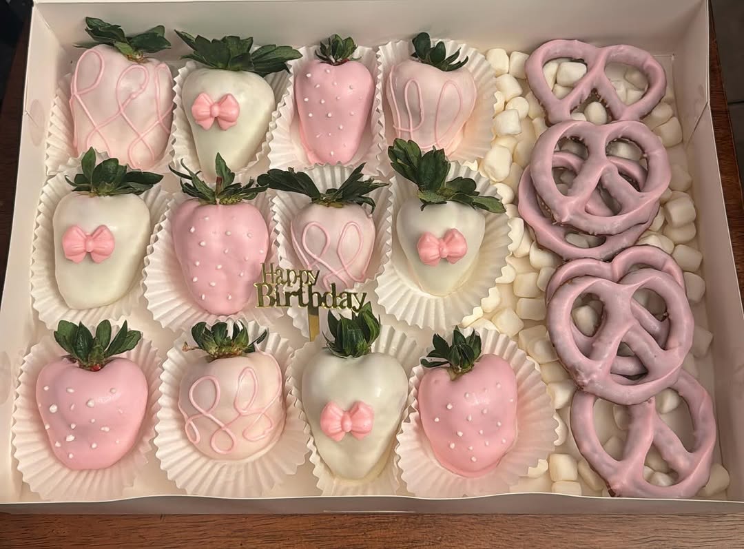 Feminine birthday box with twelve decorated strawberries in multiple pink styles including polka dots, fondant bows, and crosshatch drizzle, plus pink pretzels and a gold topper.