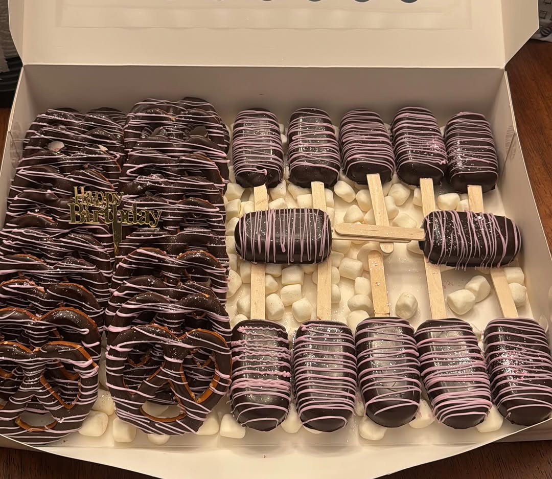 Birthday variety box of dark chocolate pretzels, cakesicles, and possibly Oreos with pink and white drizzle and a gold Happy Birthday topper.