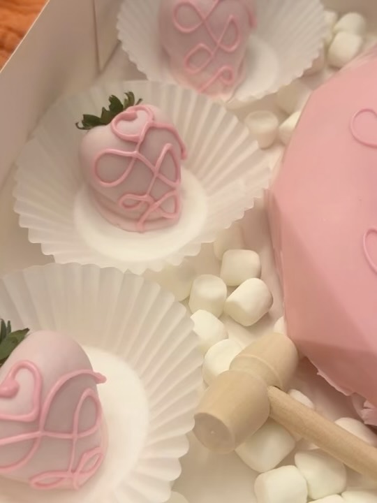 Close-up of pastel pink Valentine's strawberries with looping heart and swirl piped drizzle designs alongside a smooth pink breakable heart bomb.