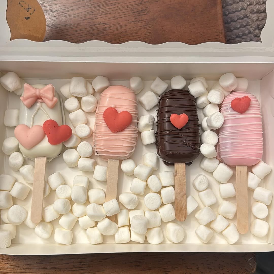 Four Valentine's cakesicles with a pink fondant bow, overlapping hearts, drizzle lines, and red fondant heart accents in marshmallows.