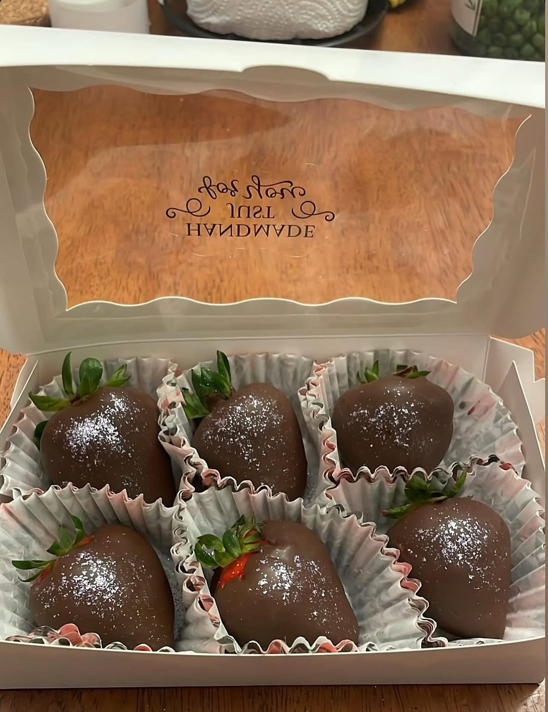 Half-dozen chocolate-covered strawberries with a light shimmer dusting in a white bakery box with a scalloped clear window lid.