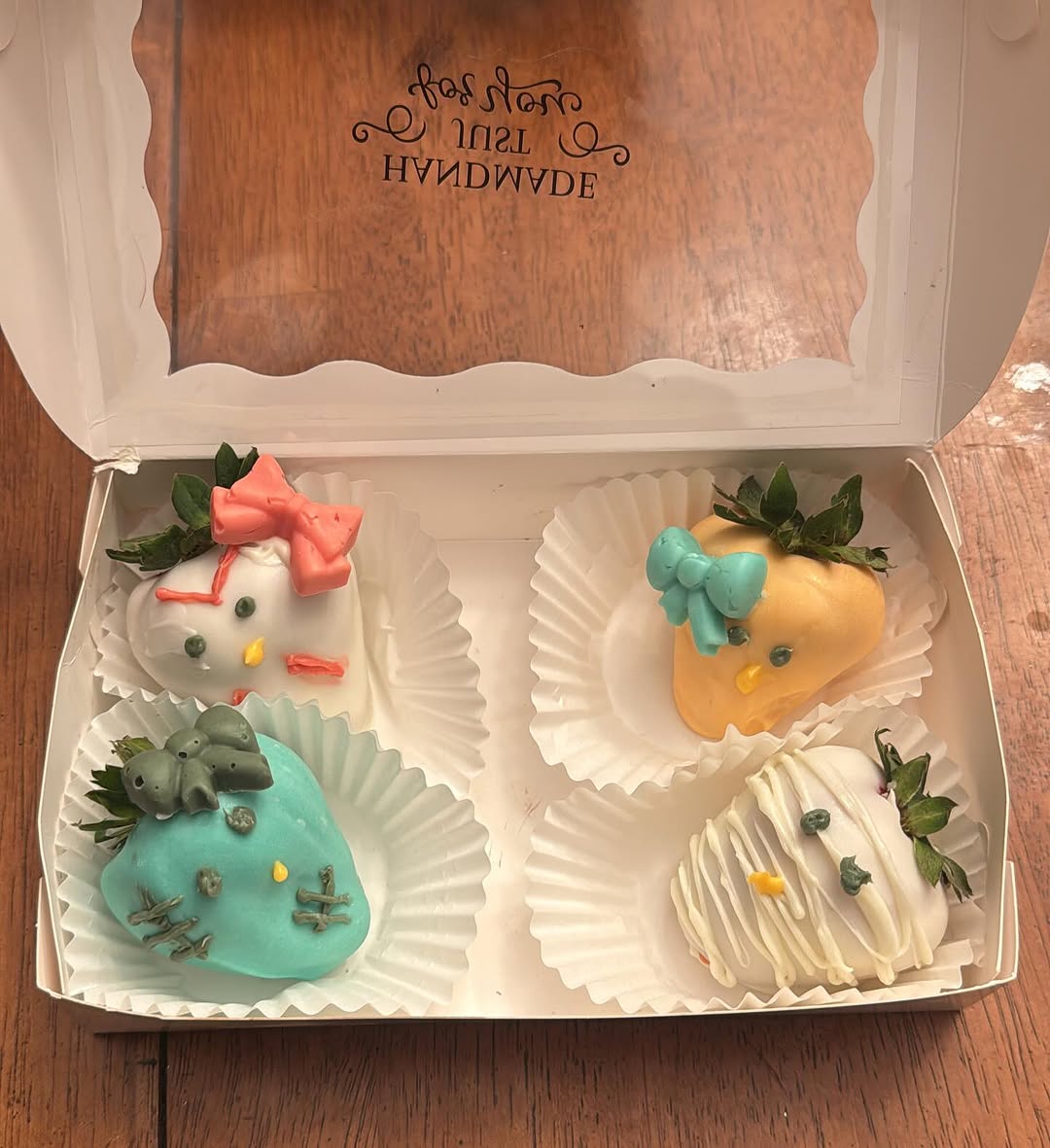 Four Sanrio character-decorated chocolate strawberries including a Hello Kitty design with pink bow and a teal character in a white bakery box.