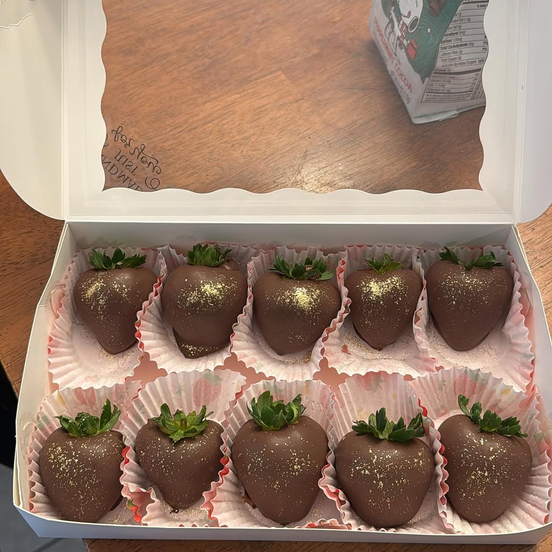 Ten dark chocolate strawberries with luxurious gold luster shimmer dust arranged in two rows inside a white bakery box with pink striped liners.